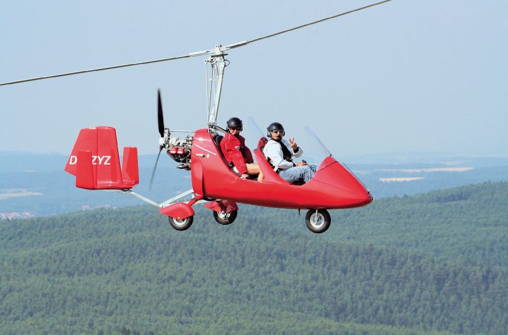 Urlaubserlebnisse: Ein kleiner roter Hubschrauber mit zwei Personen an Bord fliegt über einen Wald