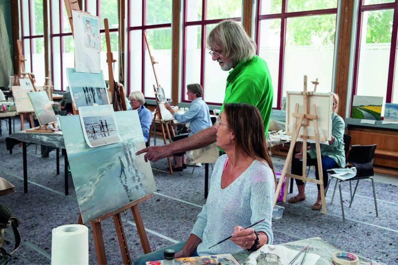 Eine Frau hält einen Pinsel in der Hand und schaut auf ein Gemälde. Ein Mann steht neben ihr und zeigt auf eine Stelle des Bildes (Urlaub und Kunst).