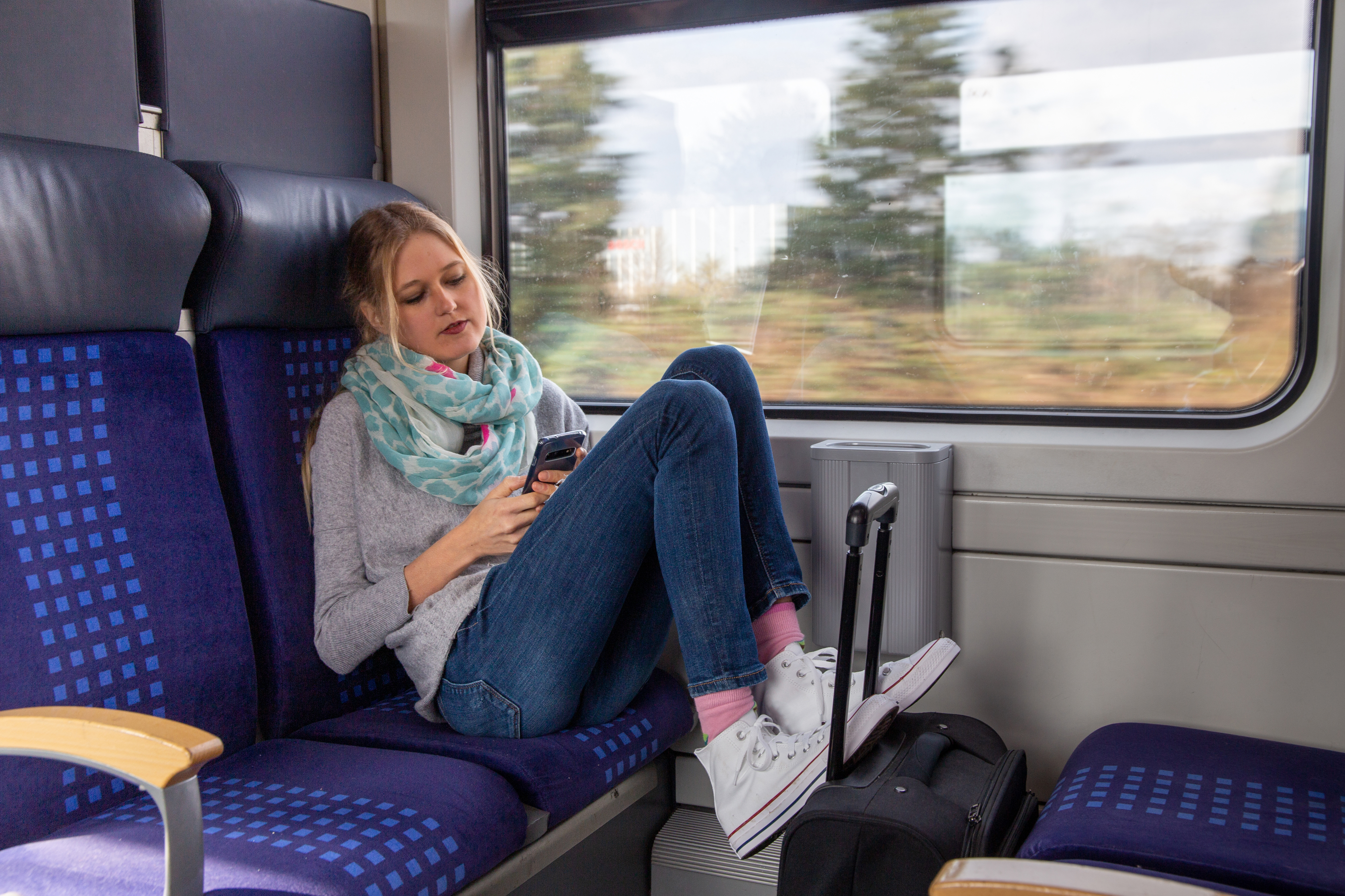 Junge Frau sitzt entspannt mit Smartphone im Zug, Füße auf dem Sitz, Koffer neben ihr.